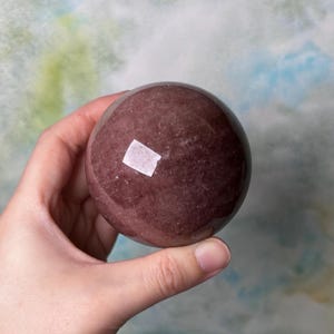 May include: A polished, round, reddish-brown crystal sphere. The sphere has a smooth surface with subtle variations in colour and texture. The crystal is held in a hand against a soft, blurred background.