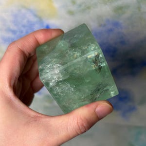 May include: A translucent green fluorite cube with visible internal fractures and natural mineral inclusions. The stone displays varying shades of light and deep green throughout its geometric structure, showcasing a smooth, polished surface texture.