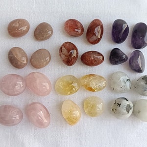 May include: A collection of 18 tumbled stones in various colours, including pink, brown, orange, yellow, white, and purple. The stones are smooth and polished, and they are arranged in rows on a white surface.