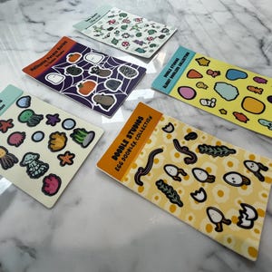May include: A collection of Doodle Studios sticker sheets featuring various designs. The sheets include themes like sea creatures, Halloween, and abstract shapes. Each sheet has a different color scheme and illustrations, with text labels indicating the collection name.