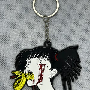 May include: Black and white enamel keychain featuring a cartoon-style illustration of a person with black hair and a snake biting their mouth. The person's eye is bleeding red.