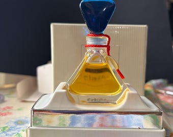 May include: A small, clear glass bottle of perfume with a blue and red stopper. The bottle is sitting on a white and silver stand. The bottle is labelled "Clarins"