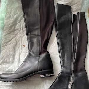 Puede incluir: Un par de botas negras hasta la rodilla. Una bota tiene una cremallera de largo completo, mientras que la otra tiene un panel elástico. Ambas botas tienen un detalle de cadena plateada alrededor de la base del tacón.