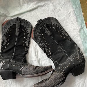 Op de afbeelding: Een paar zwarte leren cowboylaarzen met decoratieve stiksels en studs. De laarzen hebben een spitse neus en een blokhak.