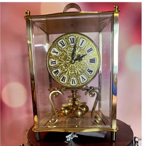 May include: A gold-toned, vintage clock encased in a glass box. The clock face is gold with black Roman numerals and ornate detailing. The pendulum features three gold spheres. The clock has a handle at the top and sits on a wooden base.