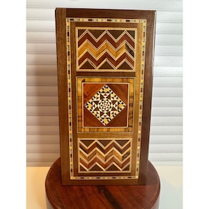 May include: A rectangular wooden box with intricate inlaid geometric patterns. The box features a dark brown frame with three panels of inlaid wood in shades of brown, tan, and cream. The central panel has a diamond pattern.