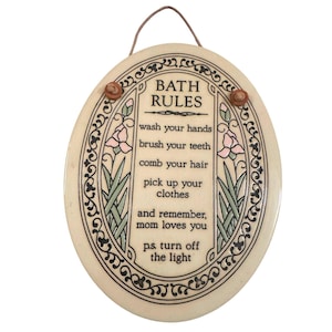 May include: Oval-shaped ceramic plaque with a cream background and a brown cord for hanging. The plaque features the words "BATH RULES" and instructions like "wash your hands" and "brush your teeth". Decorative floral and scroll elements frame the text.