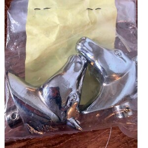 May include: Two polished chrome metal ornaments shaped like horse heads, each featuring a circular mounting base with a visible screw hole. The pair of metallic horse head figures are enclosed within a clear plastic bag with a yellow paper insert.