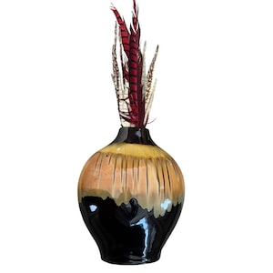 May include: A decorative ceramic vase with a black base, a tan and yellow midsection, and a black rim. The vase is filled with red, brown, and white feathers. The vase has vertical grooves and a glossy finish.