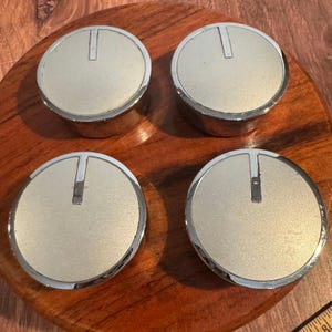 May include: Four silver and beige knobs with a metallic finish. Each knob has a circular face with a raised, rectangular indicator. The knobs are arranged on a dark wooden surface.