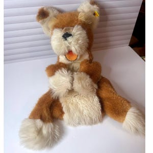 May include: A vintage plush toy dog with brown and cream fur. The dog has a fluffy face, paws, and chest, with a black nose and an open mouth revealing an orange tongue. The toy is sitting on a white surface.