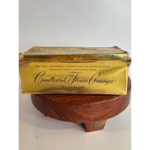 May include: A vintage gold-colored box for a candlestick flower arranger, with text that reads "Candlestick Flower Arranger" and "For a Lovely Table." The box is placed on a small, round, wooden stand.