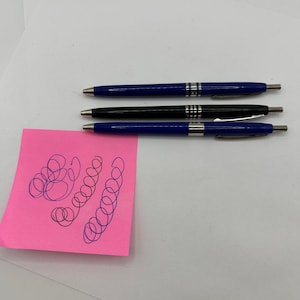 May include: Three pens: two blue and one black, with silver accents. A pink sticky note displays scribbled ink tests in blue and black. The pens are on a white surface, showcasing their design and functionality.