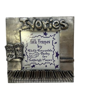 May include: A pewter-colored picture frame with the word "Ivories" at the top. The frame features a piano keyboard at the bottom and a decorative design on the left side. The center has text that reads "folk Frames by Ruth Reynolds Darby for Ashleigh Manor."