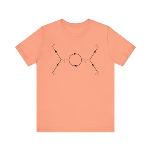 May include: A peach-colored t-shirt with a black illustration of a Feynman diagram depicting electron-positron annihilation. The diagram shows two incoming electrons and two outgoing positrons, with a photon loop in the middle.