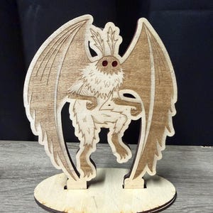 Op de afbeelding: Een houten lasergesneden sculptuur van een mot-achtig wezen met grote vleugels en rode ogen. De sculptuur is gemaakt van lichtgekleurd hout en staat op een ronde basis. Het wezen heeft een gedetailleerd ontwerp.