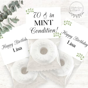 Può includere: Caramelle alla menta bianche in sacchetti di cellophane trasparente, con biglietti personalizzati che recitano "Happy Birthday Lisa" e "70 & in MINT Condition!" Sono visibili anche accenti di foglie verdi e il logo Wooden Swan Design Co.