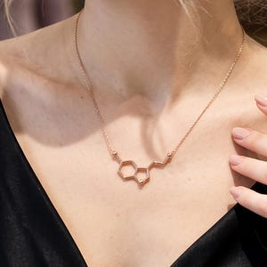 Może przedstawiać: Naszyjnik w kolorze różowego złota z zawieszką w kształcie struktury chemicznej. Zawieszka składa się z połączonych ze sobą sześciokątnych i pięciokątnych kształtów, zawieszonych na delikatnym łańcuszku spoczywającym na obojczyku.