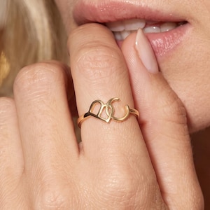 May include: A gold-toned ring featuring two interlocking letters, specifically a capital letter B and a capital letter C, connected in a continuous metal band design. The ring is worn on a finger, showcasing the polished metallic finish and the distinct shape of the two letters joined together as a single piece of jewelry.