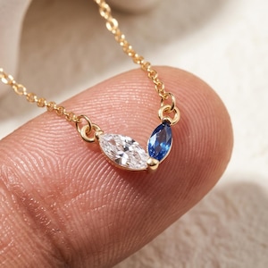 Op de afbeelding: Een delicate goudkleurige ketting met een hanger bestaande uit twee marquise-geslepen stenen. Eén steen is helder en kleurloos, terwijl de andere een diepblauwe saffierkleur heeft. De hanger hangt aan een dunne, gepolijste metalen ketting.