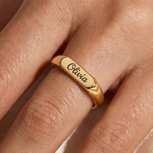 May include: A polished gold-tone band ring featuring a flat, rectangular top surface. The name Olivia is engraved in a black, cursive script font across the center of the flat surface. The ring has a smooth, rounded shank and a reflective finish.