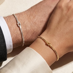 May include: A thin silver-coloured bracelet with a central knot detail worn on one wrist and a thin gold-coloured bracelet with a matching central knot detail worn on the other wrist, both featuring a smooth, minimalist metallic finish.