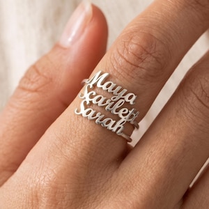 Puede incluir: Un anillo de metal plateado con tres bandas horizontales apiladas, cada una con un nombre personalizado en fuente cursiva. Los nombres Maya, Scarlett y Sarah son claramente visibles, integrados en la estructura de la banda del anillo.