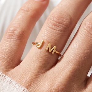 May include: A gold-toned open ring featuring two capital letters, J and M, positioned at the ends of the band. The ring has a polished metallic finish and a simple, thin circular design that wraps around the finger, showcasing the letters clearly.