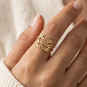 May include: Gold-colored rings with the names "Maya", "Scarlett", and "Sarah" in a delicate script. The rings are stacked on a finger, showcasing the personalized design. The rings are a piece of jewelry.