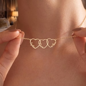 May include: A gold-toned necklace featuring three interconnected heart-shaped pendants. Each heart is outlined in a thin metal wire and incorporates a name written in a cursive script. The names Sophia, Liam, and Ava are clearly visible on the hearts.