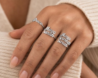Peut inclure: Trois bagues en métal argenté portées sur les doigts. La première bague porte le prénom Grace en écriture cursive. La deuxième bague affiche les prénoms Maya et Sarah superposés. La troisième bague présente Maya, Scarlett et Sarah superposés.