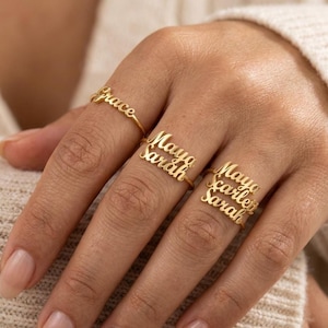 May include: Three gold-toned rings worn on fingers, each featuring custom script lettering. The first ring displays the name Grace, the second ring features the names Maya and Sarah stacked, and the third ring shows the names Maya, Scarlett, and Sarah.