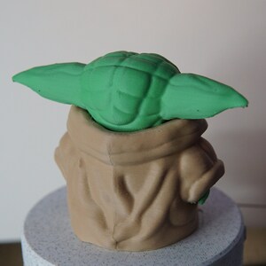 May include: A 3D-printed figurine of a character with large green ears and a tan robe. The figurine is sitting on a light gray, speckled surface. The character's head and ears are green, and the robe is tan.