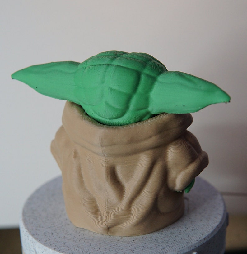 May include: A 3D-printed figurine of a character with large green ears and a tan robe. The figurine is sitting on a light gray, speckled surface. The character's head and ears are green, and the robe is tan.