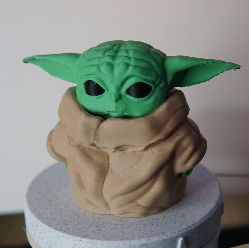 May include: A 3D-printed figurine of a green-skinned alien with large ears and black eyes, wearing a tan robe. The figure is on a round, speckled base. The character is from a popular science fiction franchise.