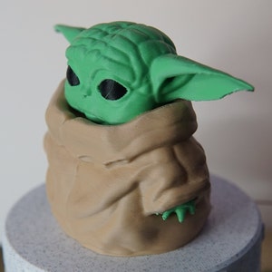 May include: A 3D-printed figurine of a green alien-like character with large ears and black eyes, nestled in a tan-colored, fabric-like pouch. The figurine is set on a light gray, textured base. The character's design is reminiscent of a popular science fiction franchise.