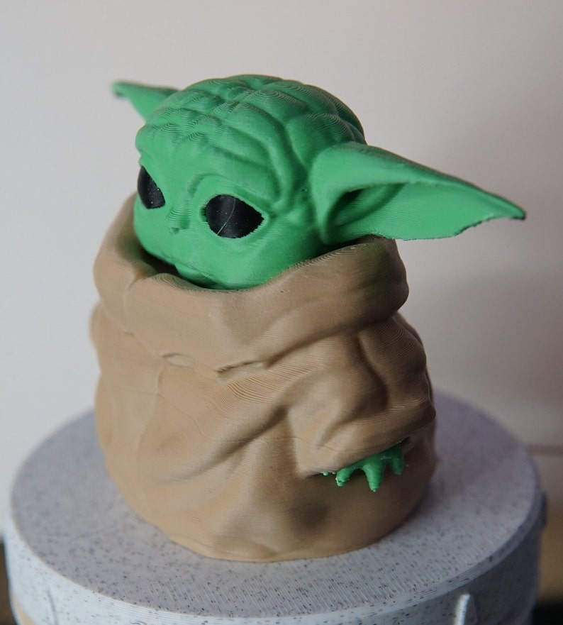 May include: A 3D-printed figurine of a green alien-like character with large ears and black eyes, nestled in a tan-colored, fabric-like pouch. The figurine is set on a light gray, textured base. The character's design is reminiscent of a popular science fiction franchise.