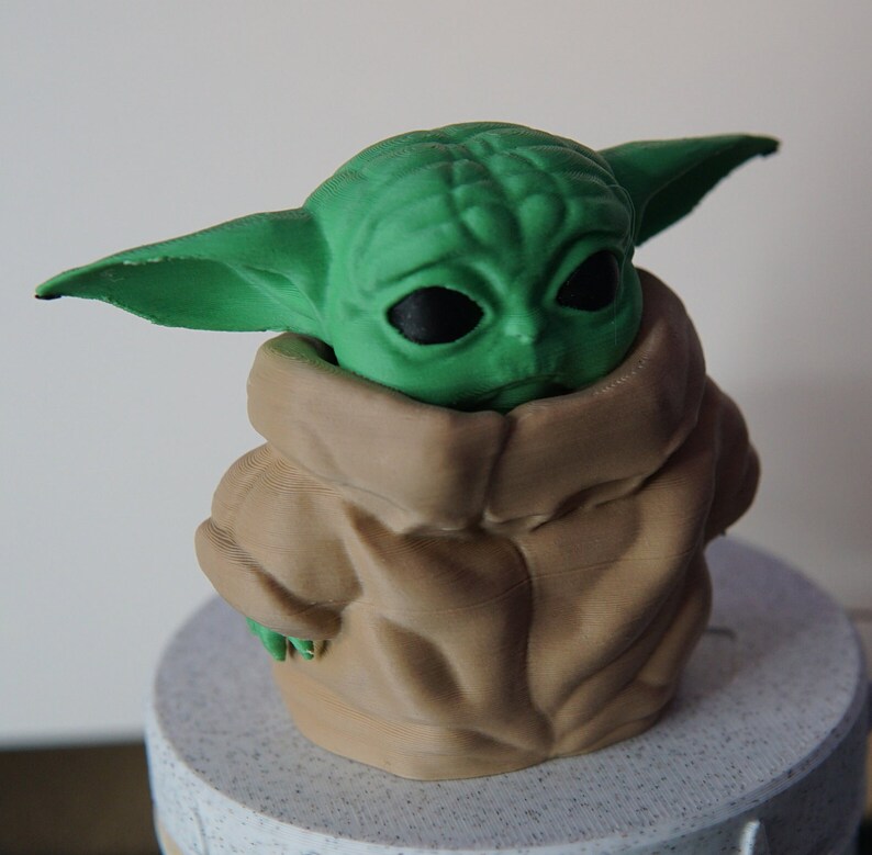 May include: A 3D-printed figurine of a character with large ears and black eyes, wearing a tan-colored cloak. The figure's head is green, and the cloak has a textured appearance. The figurine is placed on a light-colored, round base.