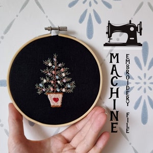 May include: A black embroidery hoop with a machine embroidery design of a potted Christmas tree with colorful ornaments. The text "MACHINE EMBROIDERY FILE" is written vertically on the right side of the hoop. The background is a white surface with a blue and gray pattern.
