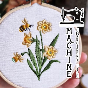 May include: Machine embroidery file of a bee and daffodils in a hoop. The design features a bee with black and yellow stripes, four yellow daffodils with green leaves, and a white background.