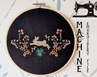 May include: A black embroidery hoop with a white rabbit jumping over a green clover, surrounded by red and white flowers and green leaves. The text "MACHINE EMBROIDERY FILE" is on the right side of the hoop. The text "BY CozyCottageStitches" is below the hoop.