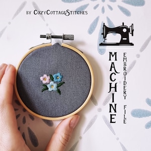 Puede incluir: Un aro de bordado gris con un pequeño diseño floral bordado en azul, rosa y blanco. El aro se sostiene en una mano. El texto "MACHINE EMBROIDERY FILE" está escrito verticalmente en el lado derecho de la imagen. El texto "BY COZYCOTTAGESTITCHES" está escrito en la parte superior de la imagen.