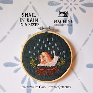 以下が含まれることがあります： 雨の中のカタツムリのデザインが施された刺繍枠。 カタツムリはオレンジとタン色の殻を持ち、白い体で、緑の葉に囲まれています。 「Snail in Rain in 6 Sizes」の文字がデザインの上に表示されています。