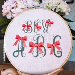 May include: Embroidered hoop art with the text "BOW" and the letters "A" and "B" in cursive with pink bows. The hoop is on a beige surface with a ladybug.