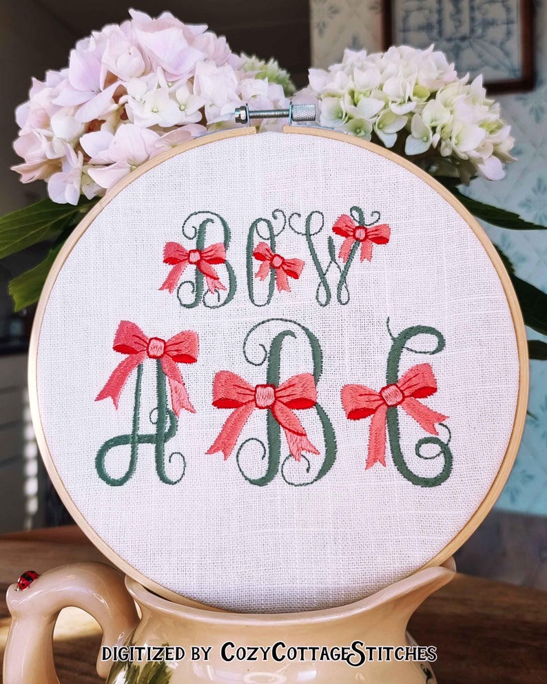 May include: Embroidered hoop art with the text "BOW" and the letters "A" and "B" in cursive with pink bows. The hoop is on a beige surface with a ladybug.