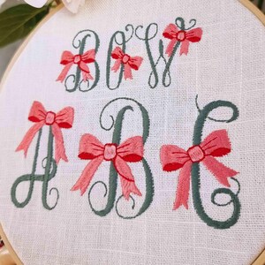 May include: Embroidered hoop art with the letters "Baby" in green cursive with pink bows. The text "Digitized by Cozy Cottage Stitches" is at the bottom of the hoop.