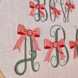 May include: Embroidered hoop art with the letters "Baby" in green with pink bows on each letter. The text "DIGITIZED BY CozyCottageSTITCHES" is at the bottom of the hoop.