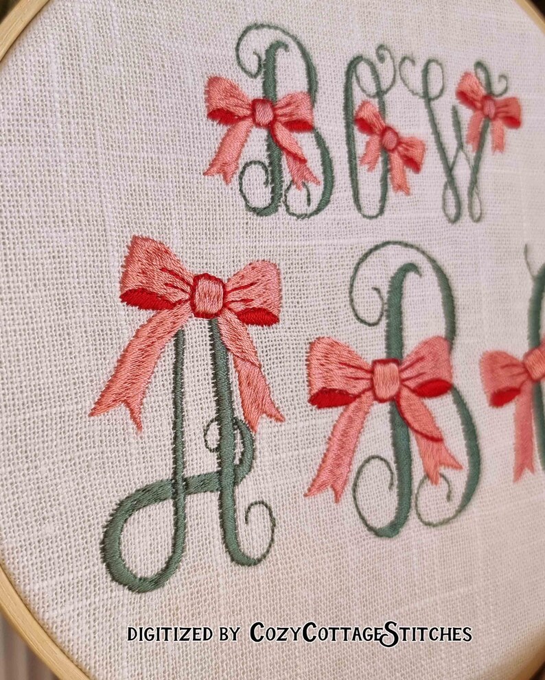 May include: Embroidered hoop art with the letters "Baby" in green with pink bows on each letter. The text "DIGITIZED BY CozyCottageSTITCHES" is at the bottom of the hoop.