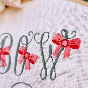 May include: Embroidered design of the word "Baby" with green lettering and pink bows. The design is on a white fabric background.