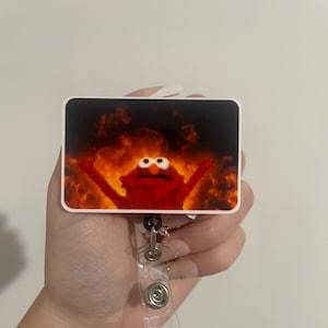 Puede incluir: Un portagafete de plástico blanco con un personaje rojo de Plaza Sésamo en llamas. El personaje tiene los brazos levantados en el aire. El portagafete está unido a un carrete retráctil de plástico transparente.